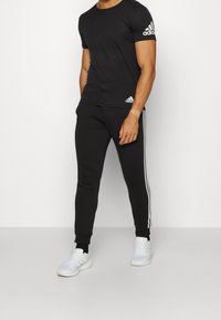 Man med svart Adidas-t-shirt och matchande svarta träningsbyxor med vita ränder, i kombination med vita Adidas-sneakers.