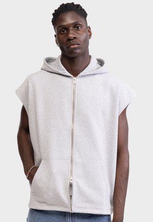 Νεαρός άνδρας με κοντές πλεξούδες, φοράει αμάνικο ανοιχτό γκρι φούτερ με φερμουάρ και μπλε τζιν, στέκεται με τα χέρια στις τσέπες μπροστά σε απλό φόντο.