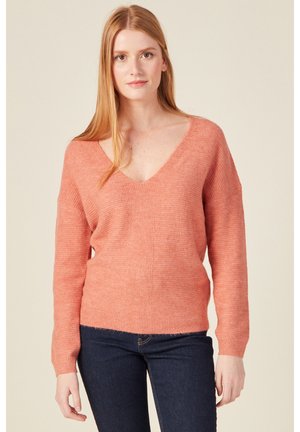 Helle korallenfarbene Strickjacke mit tiefem V-Ausschnitt, überschnittenen Schultern und gerippten Bündchen, kombiniert mit dunklen Jeans. Weiche Textur und lässige Passform.