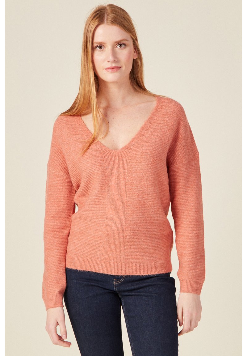 Helle korallenfarbene Strickjacke mit tiefem V-Ausschnitt, überschnittenen Schultern und gerippten Bündchen, kombiniert mit dunklen Jeans. Weiche Textur und lässige Passform.