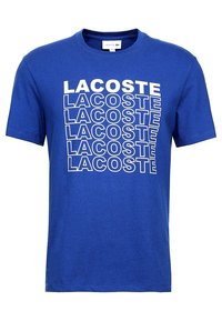 Modrá bavlněná tričko s opakujícím se bílým textem "LACOSTE". Krátké rukávy a kulatý výstřih, s hladkou, bez vzoru texturou.