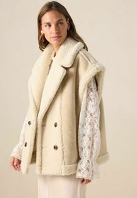 Femme portant un gilet sans manches en shearling beige sur une blouse en dentelle blanche, regardant sur le côté devant un fond beige uni.