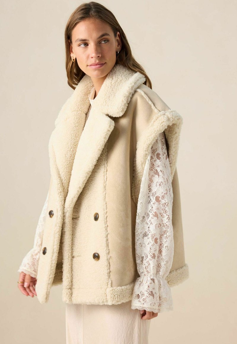 Femme portant un gilet sans manches en shearling beige sur une blouse en dentelle blanche, regardant sur le côté devant un fond beige uni.