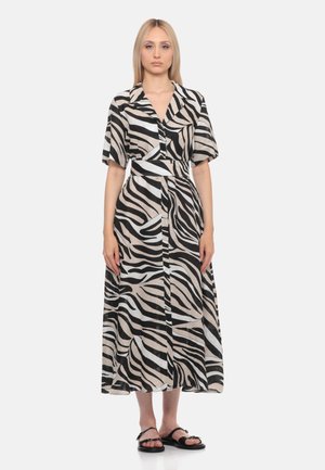 Donna bionda che indossa un vestito midi con fantasia a zebra nero, beige e bianco, maniche corte e sandali neri bassi, in piedi davanti a uno sfondo bianco.