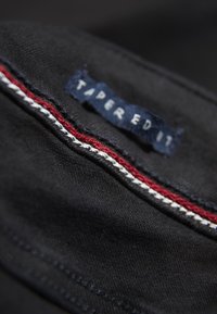 Černá tkanina s detailním záběrem na štítek "TAPER EDIT", který má červené a bílé vyšívané akcenty podél švu. Hladká textura.