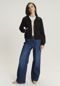 Giacca nera con zip, con polsini e vita a coste, indossata sopra un maglione a righe bianche. Abbinata a jeans a gamba larga di un blu scuro e scarpe beige.