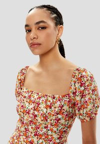 Vestido floral con escote cuadrado, mangas abullonadas y una mezcla de patrones vibrantes en naranja, rosa y crema sobre una tela ligera.