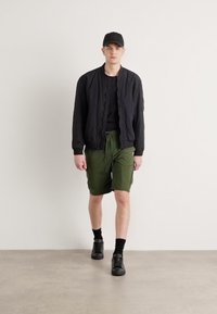 Svart bomberjakke, svart t-skjorte, grønne cargoshorts, svarte sokker og svarte sko; modellen står i en minimalistisk bakgrunn.