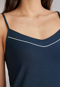 Marinblå cami med tunna axelband, med V-ringning och en vit kantdetalj över bysten. Mjuk och slät tygtextur.