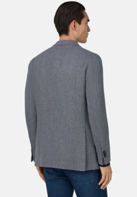 Szary żakiet w kratę o teksturowanym wzorze, z klapami, z jednym rozcięciem z tyłu i zapinanymi mankietami. Noszony na niebieskich dżinsach.