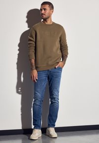 Mand med tatoveringer iført en brun sweatshirt, blå jeans og beige sneakers, der står op ad en hvid væg med skygge synlig.