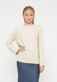 Pulover tricotat de culoare bej deschis, cu guler rotund, manșete și tiv cu dungi. Modelul este asortat cu o fustă bleu marin, având un croi lejer.