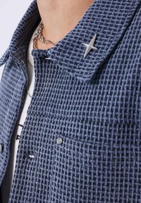 Giacca blu navy con trama intrecciata, dettaglio a stella argentata sul colletto e chiusura con bottoni, indossata sopra una camicia bianca.
