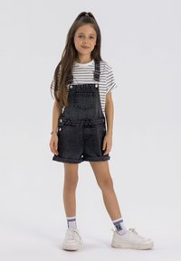 Svarta denimoveraller med rullade fållar, fästa med silverknappar, bärs över en vit och svart randig t-shirt, ihop med vita sneakers.