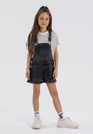 Schwarze Denim-Overall mit hochgekrempelten Säumen, gesichert mit silbernen Knöpfen, getragen über einem weiß-schwarz gestreiften T-Shirt, kombiniert mit weißen Sneakern.