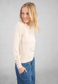 Maglione beige chiaro con collo rotondo, maniche lunghe e un'unica accentatura di bottoni ai polsini. Tessuto morbido e attillato sui fianchi.