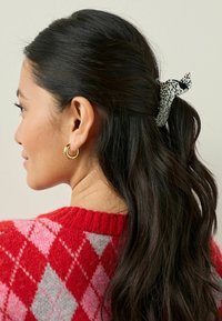 Svartvit prickig scrunchie håller tillbaka mörkt vågigt hår. Modellen bär en röd tröja med rosa och grå diamantmönster. Ett guldörhänge syns.