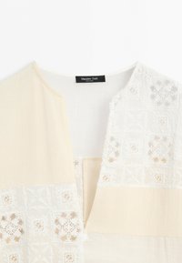 Detail béžového a bílého oděvu s texturovanou látkou a vzory z krajkových čtverců, kulatým výstřihem a štítkem Massimo Dutti.