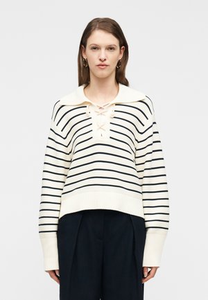 Donna che indossa un maglione a righe crema e nere con collo allacciato e polsini larghi, abbinato a pantaloni neri a gamba larga.