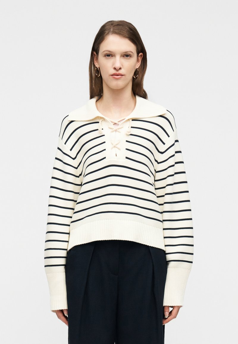 Γυναίκα που φοράει πουλόβερ πλεκτό εκρού και μαύρο με ρίγες, με δαντελωτό κορδόνι στο λαιμό και φαρδιά μανίκια, συνδυασμένο με μαύρο παντελόνι φαρδιάς γραμμής.