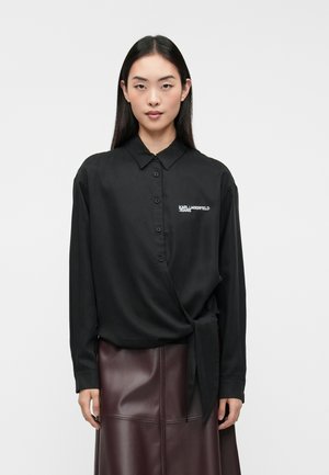 Chemise noire à manches longues et design enroulé. Présente un col et un logo blanc sur la poitrine. Texture de tissu lisse.