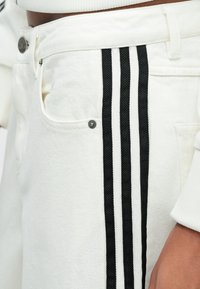 Λευκό τζιν παντελόνι με δύο μαύρες κάθετες ρίγες, μεταλλικό κουμπί και πλαϊνή τσέπη. Το ύφασμα έχει υφή με ανάγλυφο φινίρισμα.