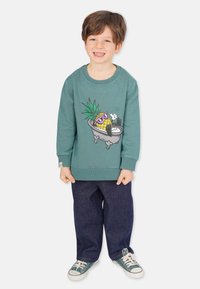 Teblås sweatshirt med en ananasgrafik, mörka denimbyxor och gröna sneakers. Har ribbade muddar och fåll.