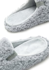 Pelēki pūkaini čības, kas izgatavotas no mīkstas, teksturētas auduma ar gludu iekšējo oderi un platu zoli. Sānos ir maza pelēka birka.