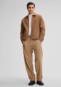 Bruine wollen zip-up jas met zijzakken, gecombineerd met beige wijde broekspijpen. Model staat tegen een neutrale achtergrond. Zwarte schoenen.