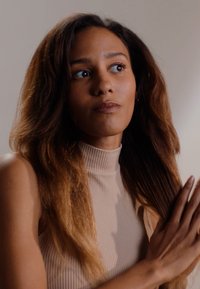 Kobieta z długimi, falującymi brązowymi włosami, ubrana w beżowy, prążkowany sweter z wysokim kołnierzem. Ma neutralny wyraz twarzy i splata ręce.