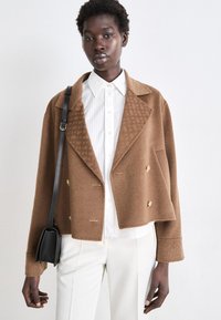 Veste cropped en mélange de laine marron avec un col cranté, un design double boutonnage, des boutons dorés et un motif texturé sur le col.