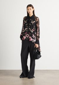 Blouse noire transparente à motif floral, manches longues et col ; assortie à un pantalon large à rayures et un petit sac à main noir.