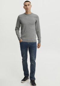 Grå strikkegenser med rund hals, sammen med blå denimjeans og svarte sneakers, stående mot en hvit bakgrunn.
