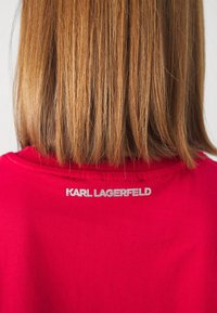 Cabello castaño claro de longitud media y lacio, sobre una camiseta roja con el texto plateado "KARL LAGERFELD" en la parte superior de la espalda.