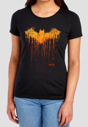 Woman wearing black t-shirt with distressed orange Batman logo dripping downward and small Batman symbol near the bottom right.