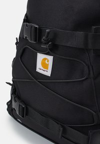 Sort stof rygsæk med justerbare stropper, hardware spænder og et hvidt mærke med et gult logo. Tekstureret overflade med snore detaljer.