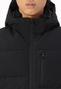 Chaqueta acolchada negra con un alto cuello, cierre de cremallera y un bolsillo con cremallera en el pecho. Textura suave y diseño ligero.