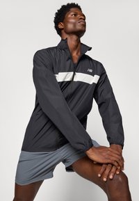 Black lightweight jacket with a white horizontal stripe and zippered collar. Paired with gray athletic shorts, showcasing a sleek design.