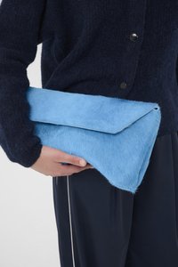 Pochette bleu clair en matière duveteuse, dotée d'un rabat incliné. Le sac est tenu dans une main et contraste avec un pull sombre.