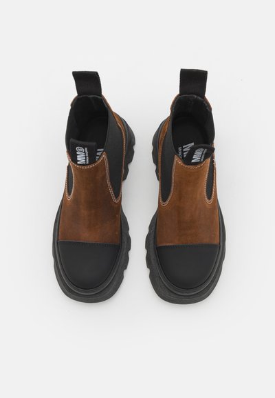 Botas de tobillo de ante marrón con punta y suela de goma negra. Presentan paneles laterales elásticos y tiradores para ponerlas y quitarlas fácilmente. Superficie texturizada.