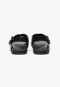Sandales à glissière en cuir noir avec une semelle plate, une sangle à boucle ajustable et un design à bout ouvert ; texture lisse et style minimaliste.