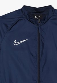 Námořnická modrá větrovka s hladkou texturou, s předním zipem a bílým logem Nike na levé části hrudníku.
