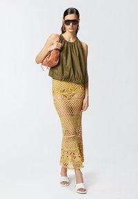 Zelena majica bez rukava s naboranim izrezom, žuta mrežasta suknja s čipkastim uzorkom, bijele sandale na klizanje i ružičasta torba za rame.