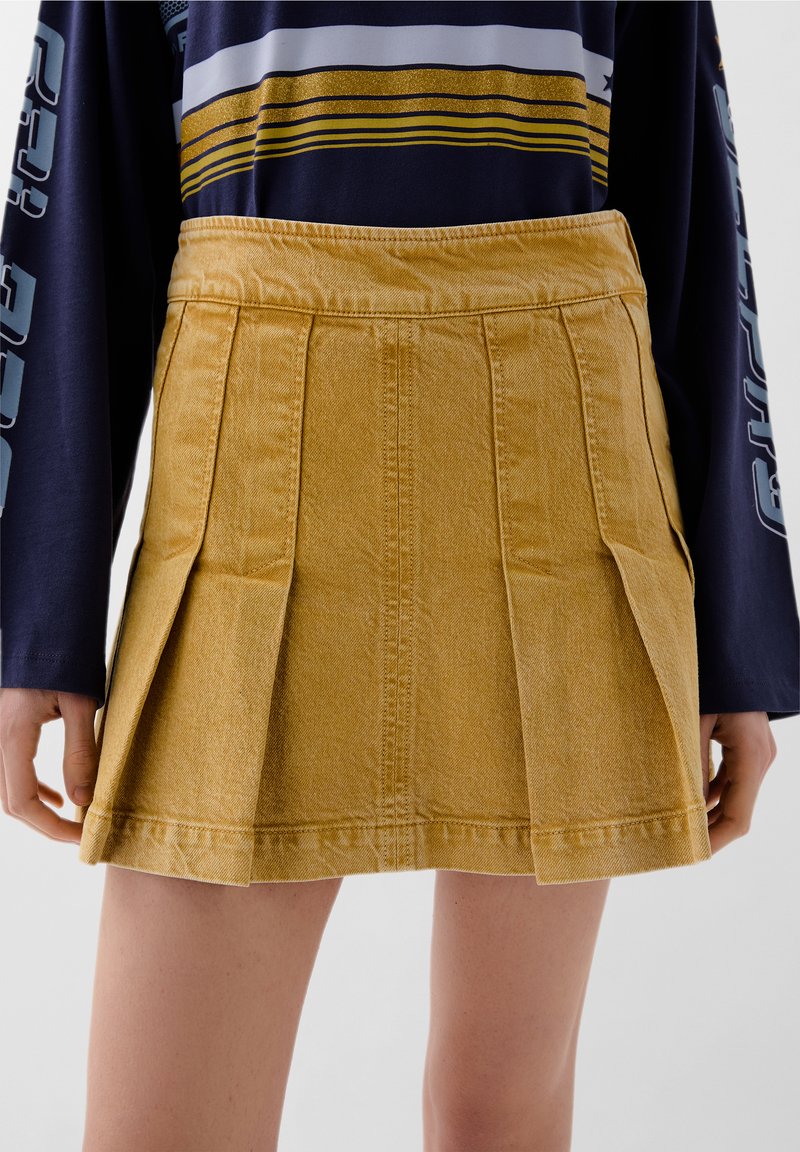 Frau in einem senfgelben, plissierten Denim-Minirock und einem marineblauen Langarmshirt mit gestreiften gelben und weißen Details.