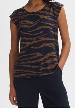 Frau trägt ärmelloses schwarzes Top mit braunem Tigerstreifenmuster und schwarze Hose, eine Hand in der Tasche.