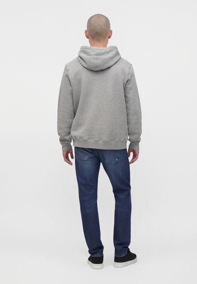Homme debout de dos, portant un sweat à capuche gris clair, un jean bleu et des baskets noires à semelles blanches.