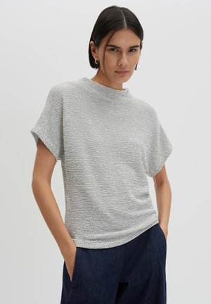 Vrouw met kort donker haar draagt een gestructureerde lichtgrijze top met korte mouwen en donkerblauwe broek, staat met handen in de zakken tegen een effen achtergrond.