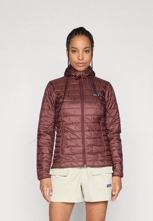 Kvinde med flettet hår iført en burgundy quiltet jakke med lynlås foran og beige cargoshorts mod en ensfarvet baggrund.