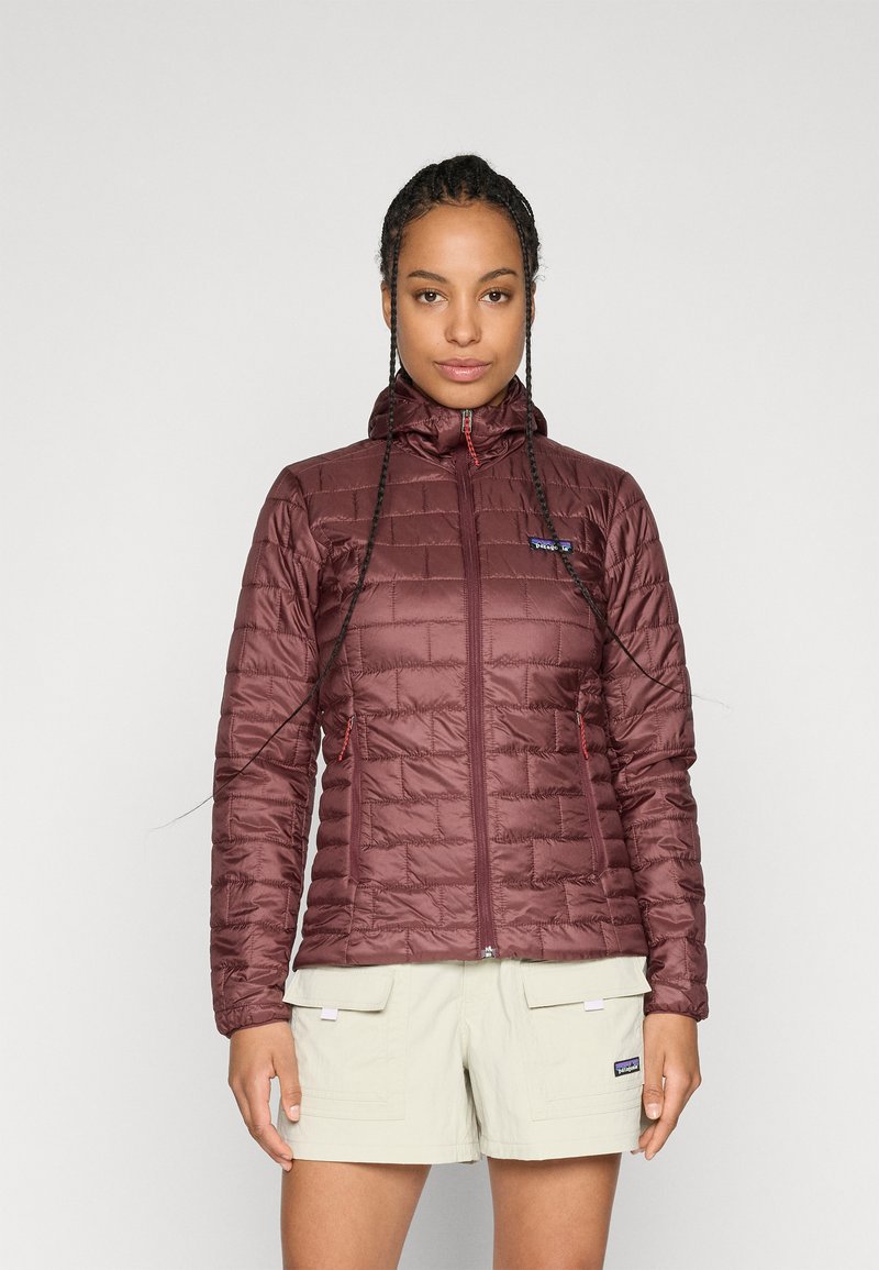 Femme avec de longues tresses portant une veste matelassée bordeaux avec capuche et un short cargo beige, se tenant devant un fond blanc uni.