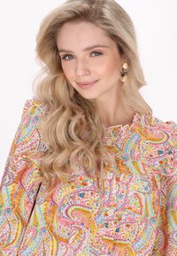 Blusa colorata con motivo paisley, caratterizzata da un colletto arricciato, dettagli con bottoni e una combinazione di tonalità di rosa, giallo, blu e arancione.